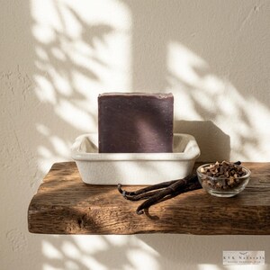 May include: A dark purple bar of soap sits in a white ceramic dish on a rustic wooden shelf. Vanilla beans and a small bowl of chopped ingredients are also on the shelf. The soap is labeled "KVK Naturals".
