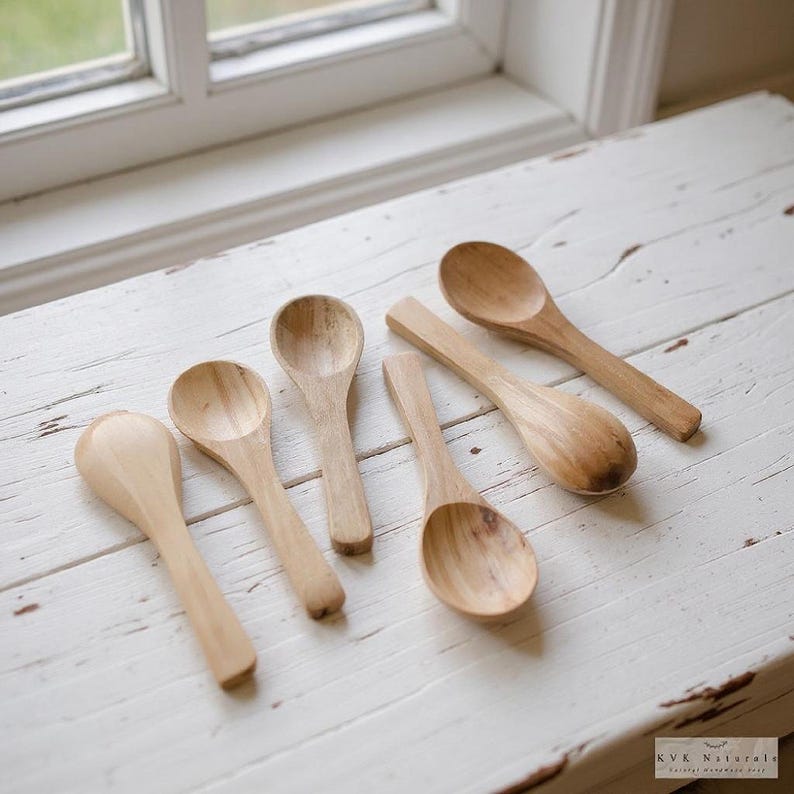 May include: Six wooden spoons of different sizes are displayed on a white, distressed wooden surface. The spoons are crafted from light-coloured wood, revealing natural grain details. These spoons are designed for kitchen use.