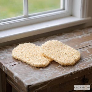 May include: Two beige, rectangular bath sponges with a textured, loofah-like appearance are displayed on a weathered wooden surface. The sponges are positioned side-by-side, suggesting they are a set. The background includes a window and a glimpse of greenery.