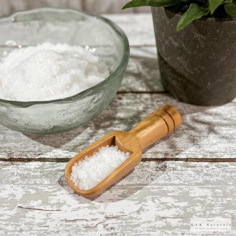 May include: A wooden scoop filled with white salt sits on a distressed white wooden surface. A clear glass bowl filled with salt is in the background, along with a potted plant. The scoop is next to the bowl.