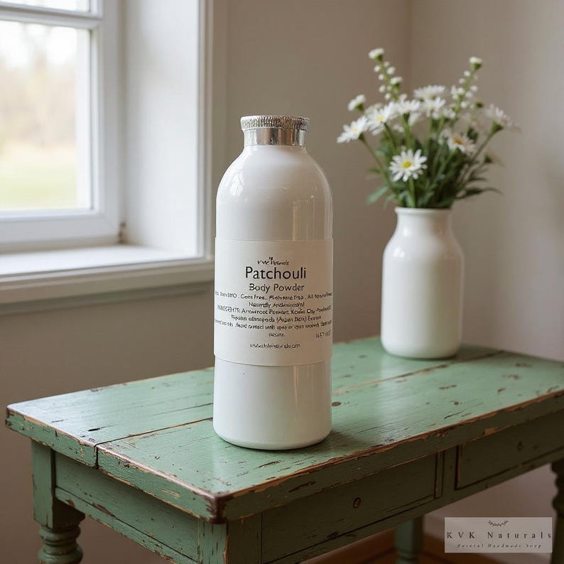 May include: White body powder bottle with a silver lid and a label that reads "Patchouli Body Powder." The bottle sits on a distressed green table next to a white vase with white flowers. The background includes a window.