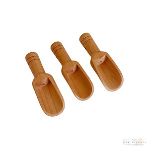 May include: Three small, light-brown wooden scoops with rounded bowls and short handles. Each handle has three carved horizontal lines near the top. The scoops are arranged diagonally against a white background, suggesting a natural or rustic theme.