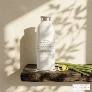 May include: A white bottle of "Lemon Grass Body Powder" with a silver cap sits on a wooden shelf. The label includes text and a logo. Fresh lemongrass and a lemon peel are arranged on the shelf. The background is a neutral wall with shadows.