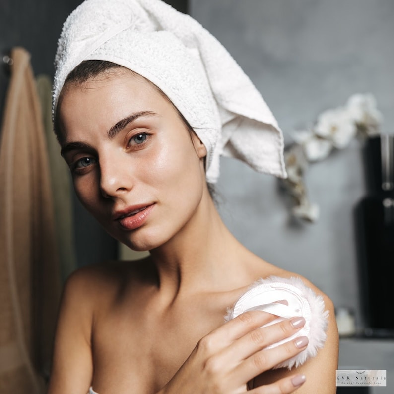 May include: A woman with a white towel wrapped around her head is holding a powder puff. The woman is in a bathroom setting. The powder puff is white and fluffy. The woman is looking at the camera.