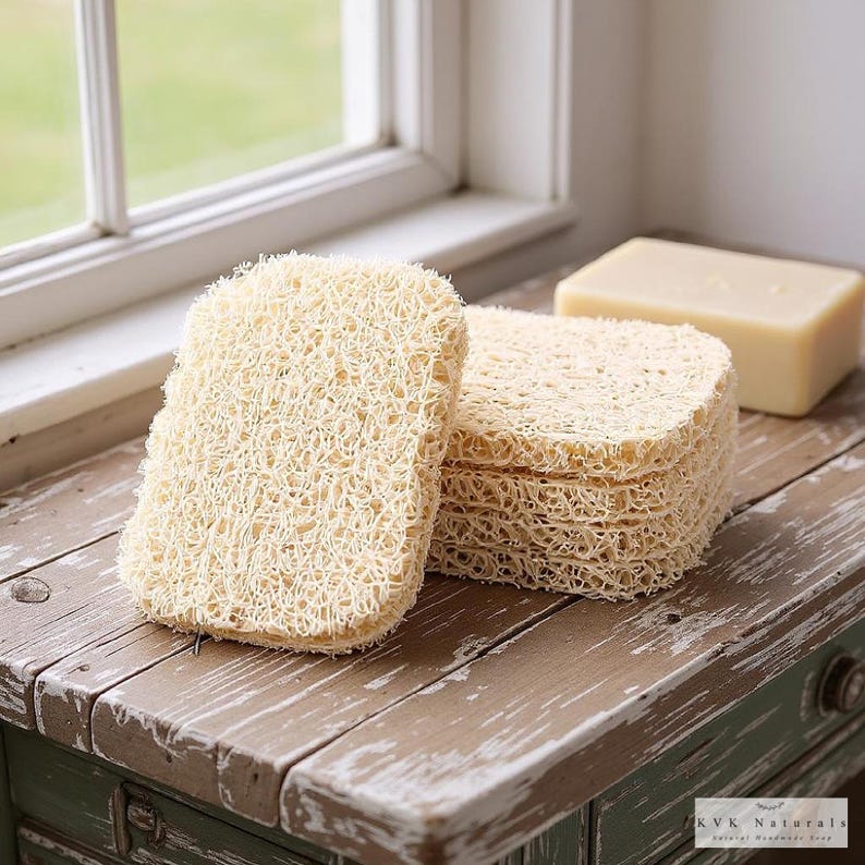 May include: A stack of beige, rectangular loofah sponges next to a bar of soap. The loofahs have a textured, woven appearance. The soap is a light beige color. The items are on a distressed wooden surface.