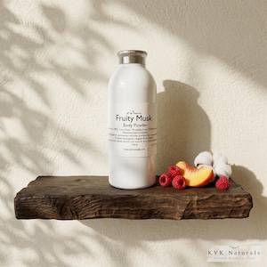 May include: A white bottle of "Fruity Musk" body powder with a silver lid sits on a rustic wooden shelf. Fresh raspberries, a peach slice, and cotton buds are arranged on the shelf. The background is a neutral wall.
