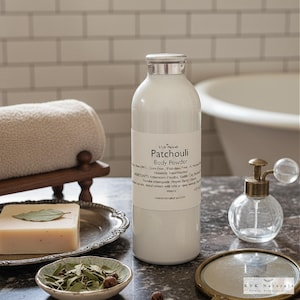 May include: A white bottle of "Patchouli Body Powder" with a silver cap sits on a marble surface. Beside it are a bar of soap with a leaf, a small bowl of herbs, a rolled towel, and a vintage perfume bottle.