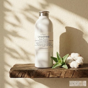 May include: A white bottle of "Unscented Body Powder" with silver cap, sitting on a rustic wooden shelf. The label lists ingredients. Beside it are an aloe vera plant and cotton. The background is a neutral wall.