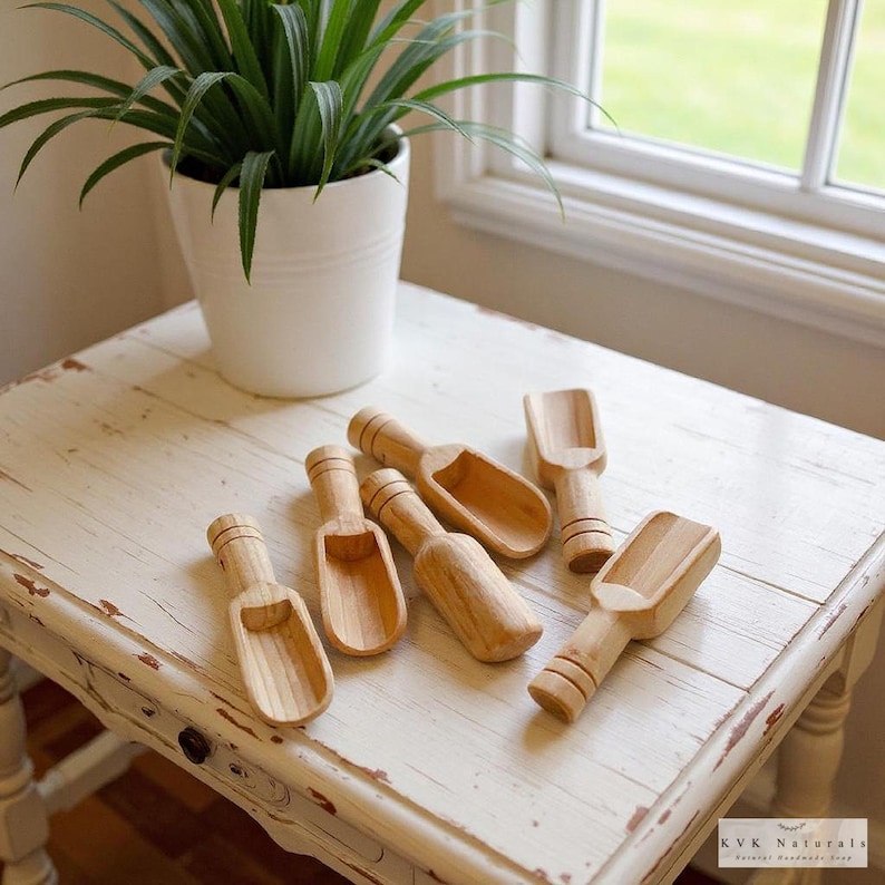 May include: A collection of small, wooden scoops with rounded bowls and short handles. The scoops are made of light-colored wood and are arranged on a distressed white table. A potted plant sits in the background near a window.