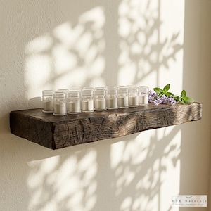 May include: A rustic, dark brown wooden shelf displays small clear jars filled with a light-colored powder. The shelf is adorned with sprigs of lavender and mint. The shelf is mounted on a light-colored wall, with shadows cast across it.