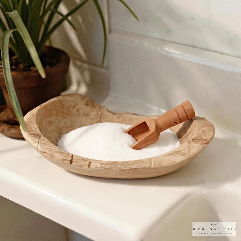 May include: A wooden bowl filled with white powder, accompanied by a small wooden scoop. The bowl has a natural wood grain finish. The scene is set in a bathroom, with a plant in the background.