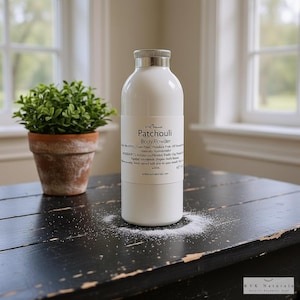 May include: A white body powder bottle with a silver cap and the text "Patchouli Body Powder." A small potted plant sits to the left. The bottle is on a black wooden surface with powder sprinkled around it.