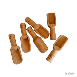 May include: Six small, wooden scoops with a light brown finish are scattered on a white background. Each scoop has a rounded bowl and a handle with two thin, dark brown bands. The scoops are likely for measuring ingredients.