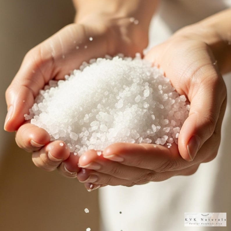 May include: A pair of hands cupping a generous quantity of white, granular salt. The salt is spilling from the hands. The image is well-lit, emphasizing the texture and purity of the salt. The product is from KVK Naturals.
