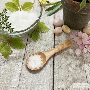 May include: A wooden spoon holds white salt crystals on a weathered wooden surface. A glass bowl contains more salt, with green leaves and thyme sprigs. Pink and yellow stones and pink flower petals are also present.