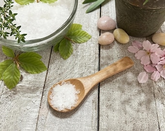 May include: A wooden spoon filled with white salt crystals rests on a weathered wooden surface. A clear glass bowl holds more salt, surrounded by green leaves and sprigs of thyme. Pink and yellow stones and pink flower petals add color.