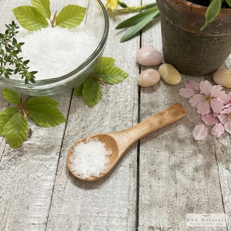 May include: A wooden spoon holds white salt crystals on a weathered wooden surface. A glass bowl contains more salt, with green leaves and thyme sprigs. Pink and yellow stones and pink flower petals are also present.