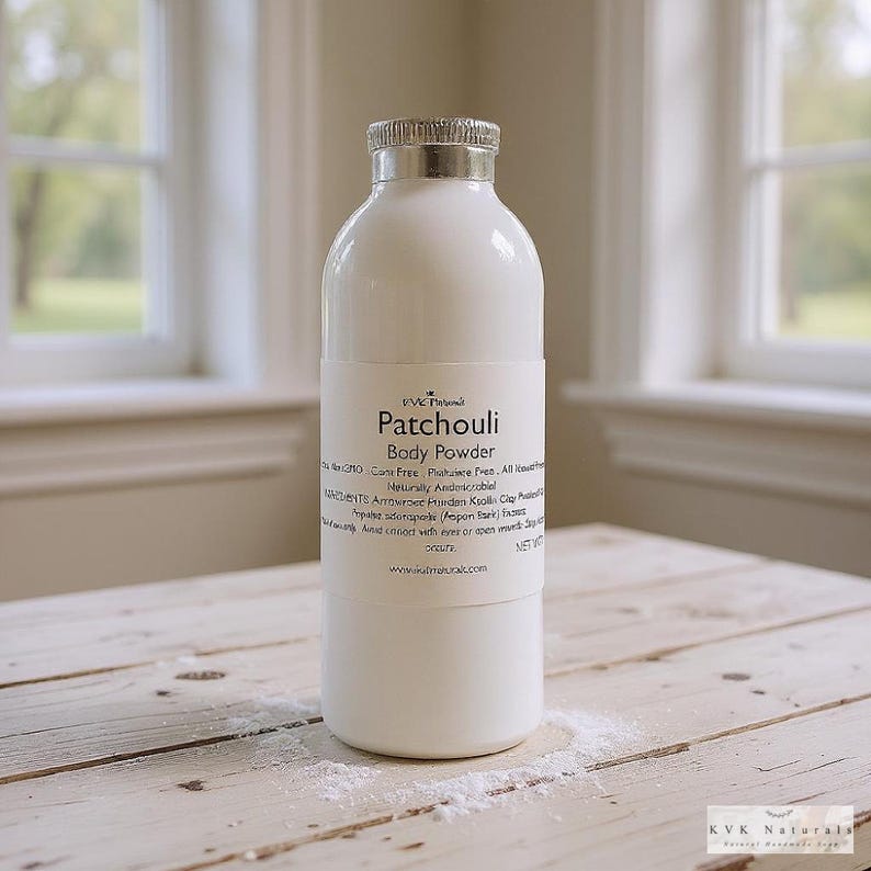 May include: A white body powder bottle with a silver cap and a label that reads "Patchouli Body Powder." The bottle is on a wooden surface with a scattering of powder. The background includes a window.