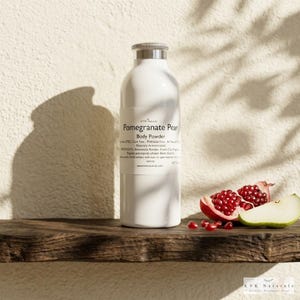 May include: A white body powder bottle with a silver cap, labeled "Pomegranate Pear." Beside it, a halved pomegranate and a slice of pear rest on a wooden surface. The background is a textured wall with shadows.