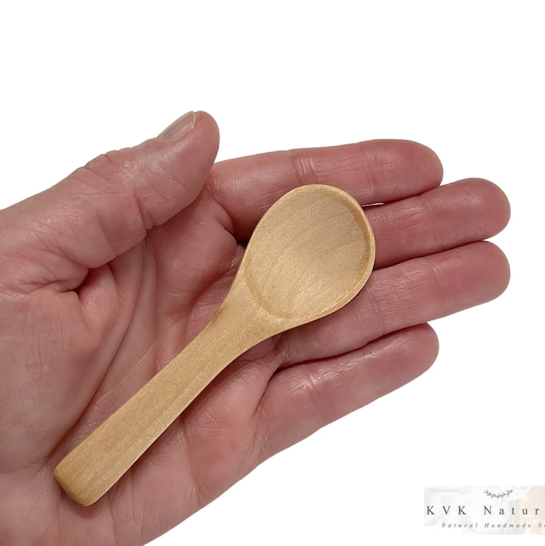May include: A small, light-coloured wooden spoon with a rounded bowl and a slender handle. The spoon is held in a hand against a white background. The spoon is approximately 10 cm long.