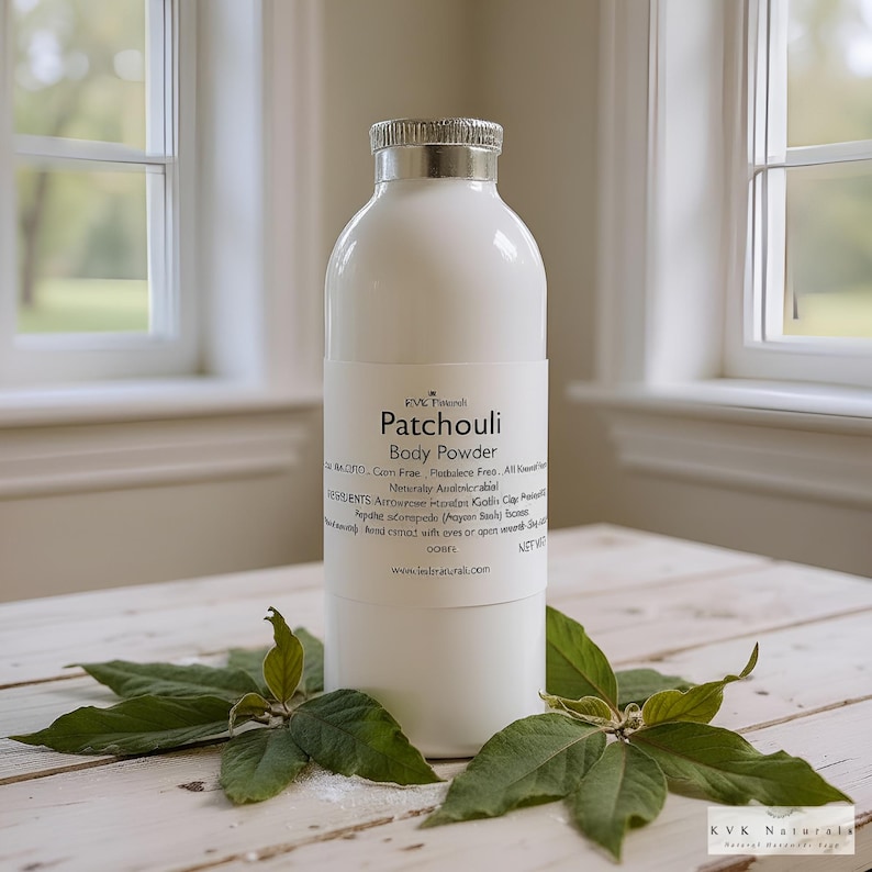 May include: A white bottle of Patchouli Body Powder with a silver lid and a label. The label includes the product name and ingredients. Green leaves are arranged around the bottle on a light-colored wooden surface.
