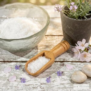 May include: A wooden scoop filled with white salt sits on a distressed white wooden surface. A clear glass bowl filled with salt is in the background. A small potted plant with lavender and white flowers and small stones are also present.