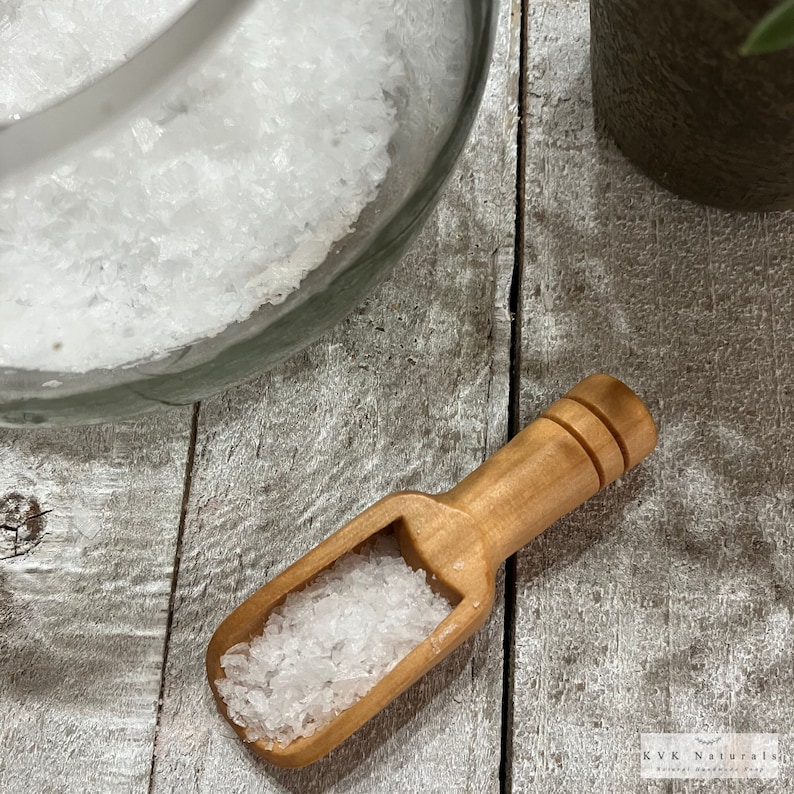 May include: A wooden scoop filled with coarse white salt sits on a weathered wooden surface. A glass jar filled with salt is in the background. The scoop is light brown and has a small handle. The image is well-lit, highlighting the texture of the salt.