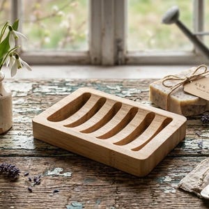 May include: A wooden soap dish with curved grooves, a bar of soap with a tag, and a small vase of white flowers sit on a rustic wooden surface. A silver spoon and a sprig of lavender are also present, creating a natural, spa-like aesthetic.