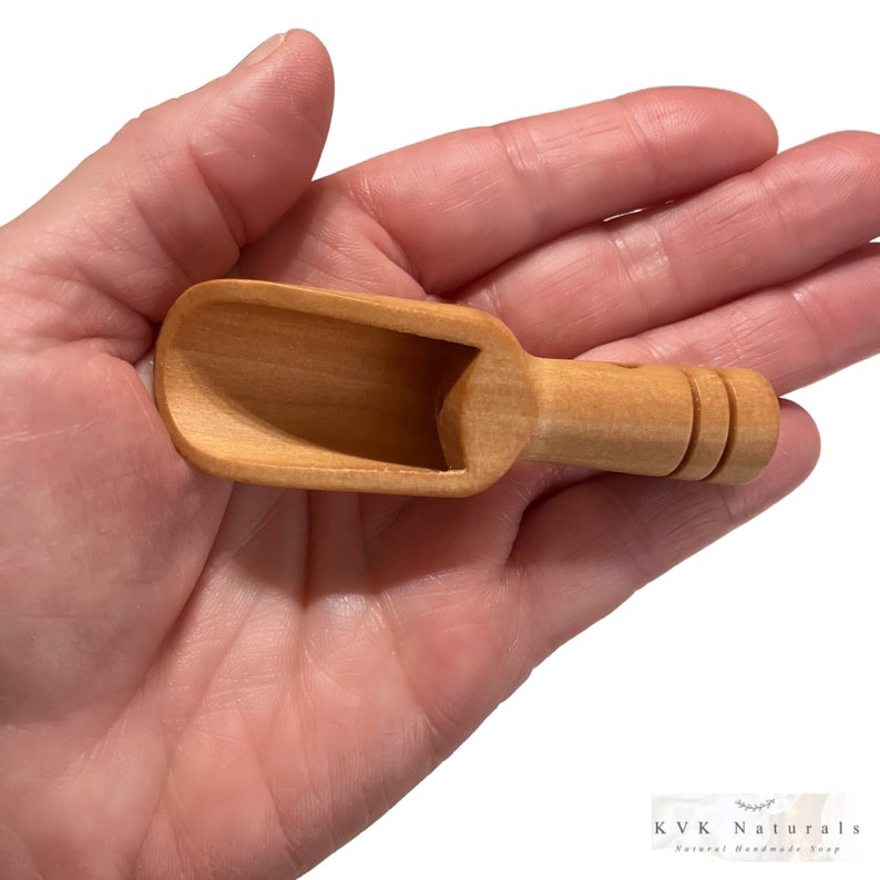 May include: A small, light-colored wooden scoop with a rounded bowl and a handle with two carved rings. The scoop is held in a hand, and the KVK Naturals logo is visible in the bottom right corner.