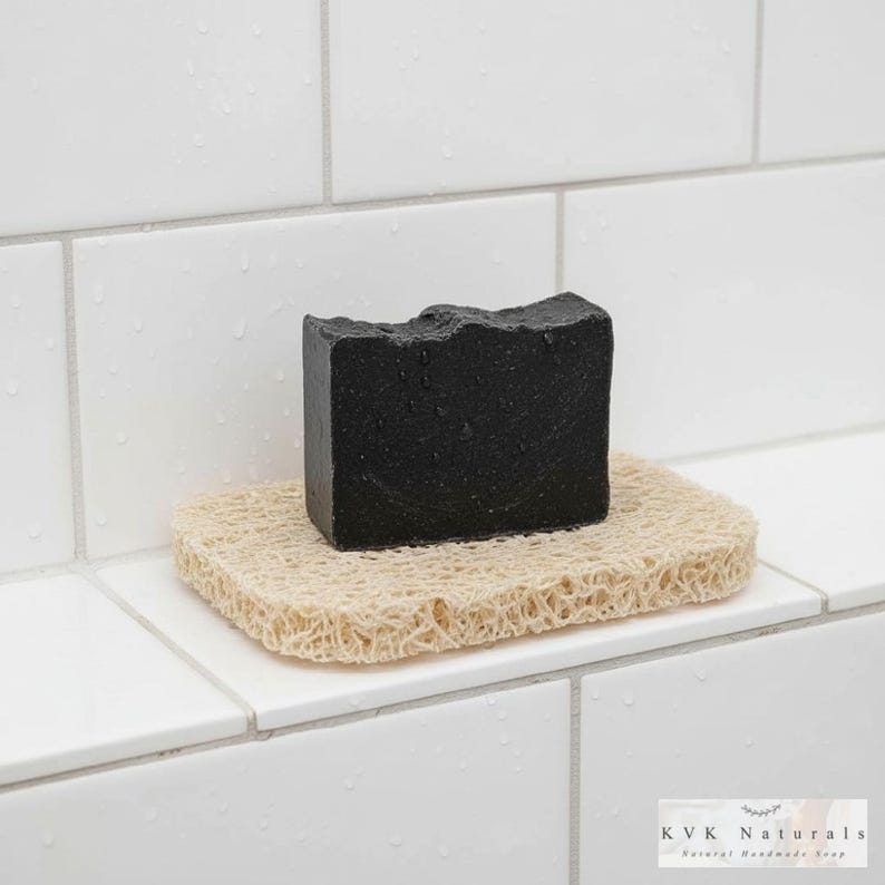 May include: A black bar of soap rests on a beige, textured soap dish. The soap has a rough, uneven top surface. The soap dish is square and made of a natural, woven material. The background is white tile.