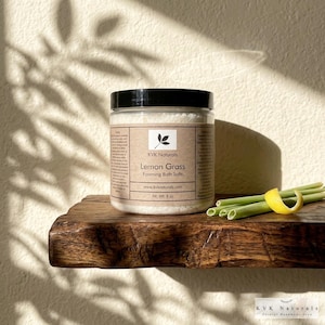 May include: A clear jar of KVK Naturals Lemon Grass Foaming Bath Salts with a black lid. The label features a leaf design and the text "Lemon Grass Foaming Bath Salts". The jar sits on a wooden shelf with lemongrass and a lemon peel.