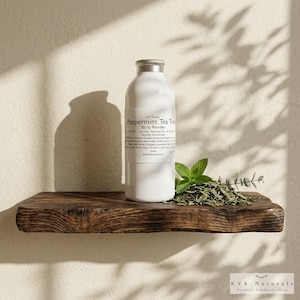 May include: A white bottle with a silver cap labeled "Peppermint Tea Tree Body Powder" sits on a rustic wooden shelf. Fresh mint leaves and dried tea leaves are arranged near the bottle. The shelf is against a beige wall.