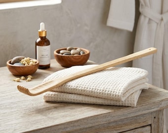 May include: A long wooden back scratcher with a curved, multi-pronged head resting on a folded white waffle-weave towel. Nearby are a small wooden bowl containing smooth grey stones and dried flowers, and a brown glass bottle with a white dropper cap.