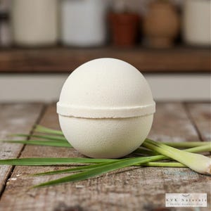May include: A light green bath bomb with a textured surface and a band around the middle sits on a wooden surface with green grass. The bath bomb is round and has a natural, handmade appearance. The bottom right corner has the text "KVK Naturals".