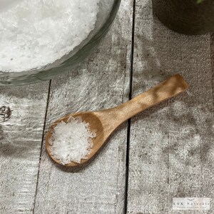 May include: A wooden spoon filled with coarse white salt rests on a weathered wooden surface. A glass jar filled with salt is in the background. The spoon is light brown, and the salt crystals are translucent.