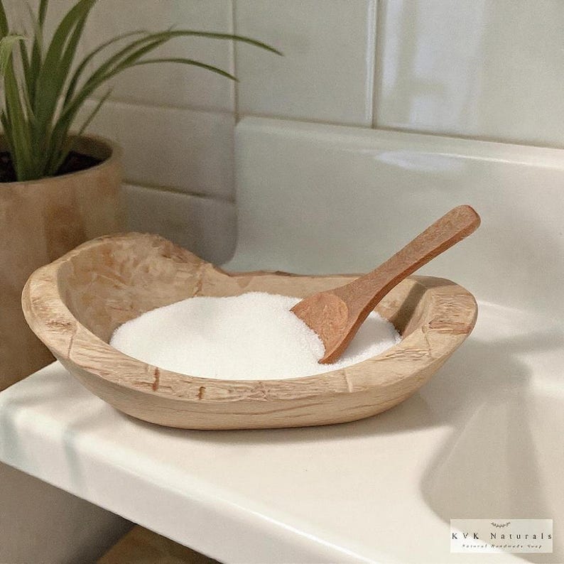 May include: A wooden bowl filled with white powder and a small wooden scoop. The bowl has a rustic, carved design. The scene is set in a bathroom or spa-like environment, with a plant in the background.