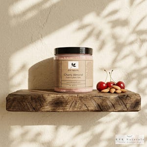 May include: A clear jar of pink Cherry Almond Foaming Bath Salts with a black lid, sitting on a rustic wooden shelf. The jar has a brown label with text and a leaf logo. Fresh cherries and almonds are arranged on the shelf. The jar contains 9 oz of bath salts.