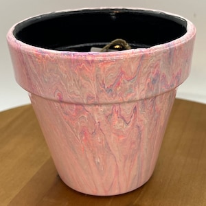 May include: A pink terracotta planter with a marbled design in shades of pink, white, and gray. The planter has a black rim and is painted with a swirling pattern.
