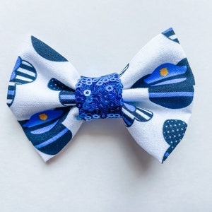 May include: A fabric bow tie with a white background and a pattern of blue and navy blue police hats. The center of the bow is embellished with blue sequins.