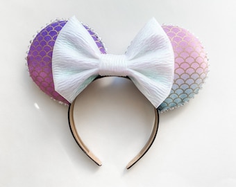 May include: A pair of mermaid-themed mouse ears with a white bow. The ears are decorated with a purple, gold, and blue scale pattern and are trimmed with small, clear beads. The headband is gold-toned.
