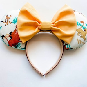 May include: A pair of mouse ears featuring a large, light orange bow. The ears are white with a repeating pattern of cartoon characters, including Timon, Pumbaa, and Simba. The headband is brown.