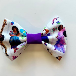 May include: A fabric hair bow featuring a purple center and a white patterned design. The pattern includes illustrations of characters in colorful dresses, flowers, and a giraffe print.