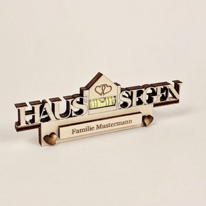 May include: A wooden sign with the German words "HAUS SEGEN" and a small house shape with two hearts. It includes a level and a space for a name, "Familie Mustermann". The sign is made of light-colored wood with a natural finish.