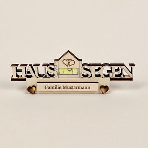 May include: A wooden house-shaped decoration with the German words "HAUS SEGEN" and "Familie Mustermann" engraved. The design includes a spirit level and heart-shaped cutouts. The wood has a natural, light brown color.