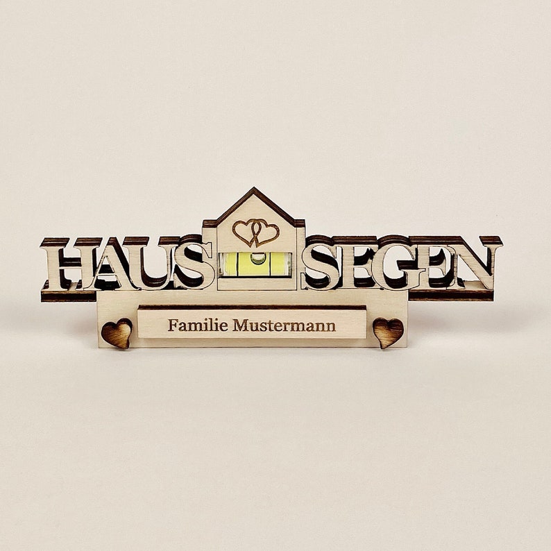 May include: A wooden house-shaped decoration with the German words "HAUS SEGEN" and "Familie Mustermann" engraved. The design includes a spirit level and heart-shaped cutouts. The wood has a natural, light brown color.