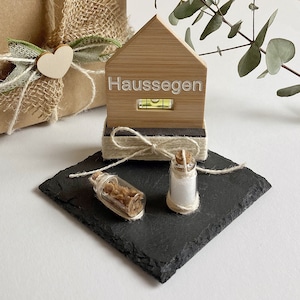 Könnte beinhalten: Ein kleines Holzhaus mit der Aufschrift "Haus Segen", einer Wasserwaage und zwei kleinen Glasflaschen mit Korkverschlüssen, alles auf einer schwarzen Schieferplatte.