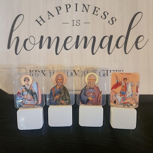 Peut inclure: Quatre petites veilleuses blanches avec des couvercles en plastique transparent. Chaque couvercle présente une image d'icône religieuse différente. Les icônes sont Saint George, Saint Nicolas, Saint Spyridon et un ange.
