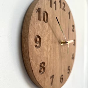 Peut inclure: Une horloge ronde en bois avec des chiffres sculptés. Le cadran de l'horloge est de couleur marron clair, avec des aiguilles dorées. L'horloge est fixée sur un mur blanc. Les chiffres sont gravés dans le bois.