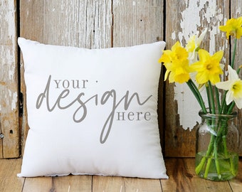 May include: A white square pillow with the text "your design here" in gray script. A vase of yellow and white daffodils sits to the right. The background is a weathered wooden wall and floor.