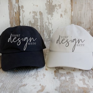 May include: Two blank baseball caps, one black and one white, are lying on a weathered wooden surface. The caps have the text "Your design here" written on them.